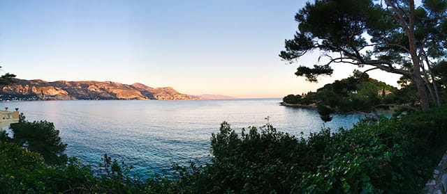 Coastal landscape with calm sea, cliffs, and trees at sunset.