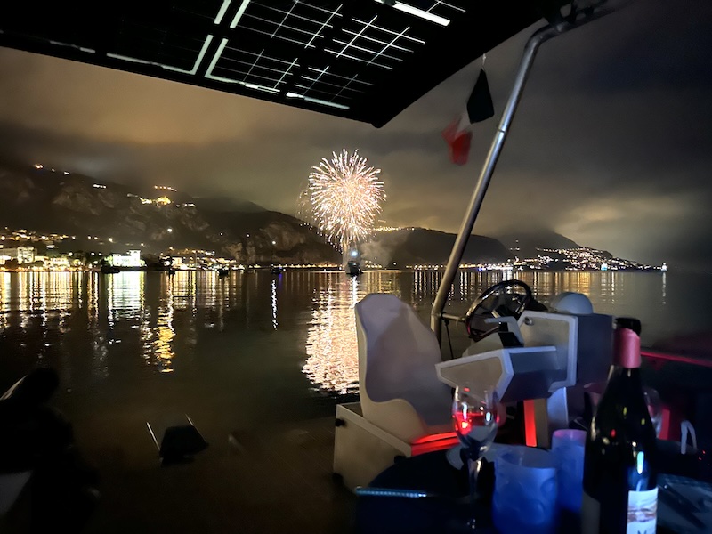 Fireworks over a calm bay at night, viewed from a boat with wine glasses.