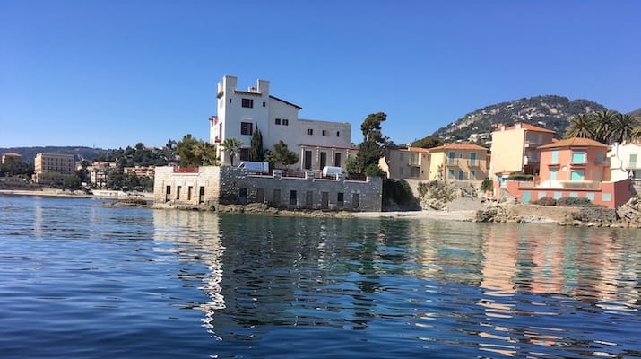 Seaside buildings with clear blue water and mountains in the background.