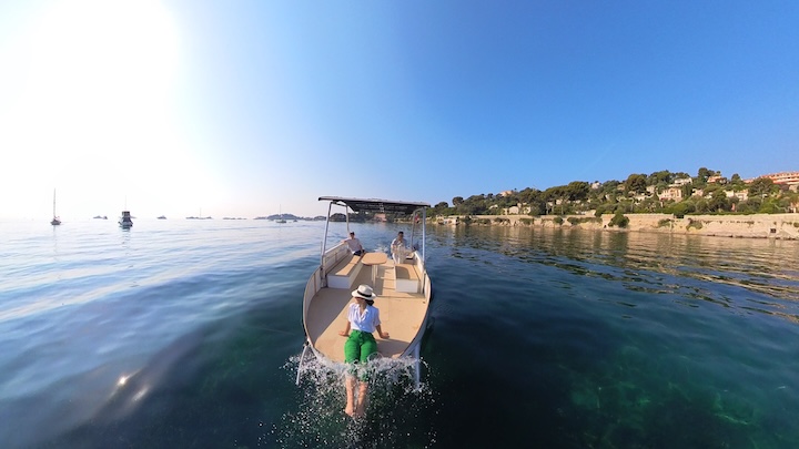 Person in green shorts standing on boat on calm sea near coastal town.