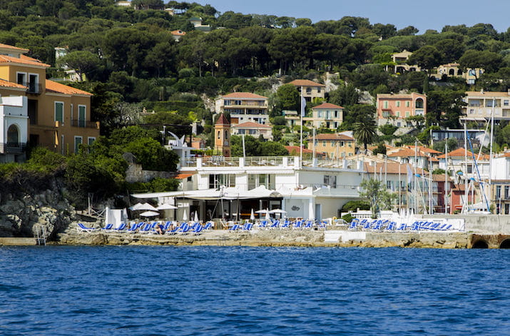 Coastal town with colorful houses, trees, and small marina by blue sea.