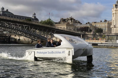 Small white boat named 'Prototype SeaBubbles #1' on a river with buildings and a bridge in the background.