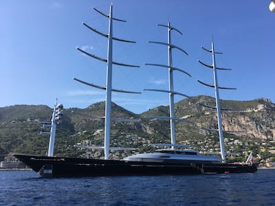 Large, modern sailing yacht with tall masts against a mountainous coastline.