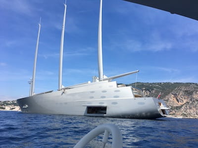 Large modern sailboat with three masts, on blue sea near steep cliff.