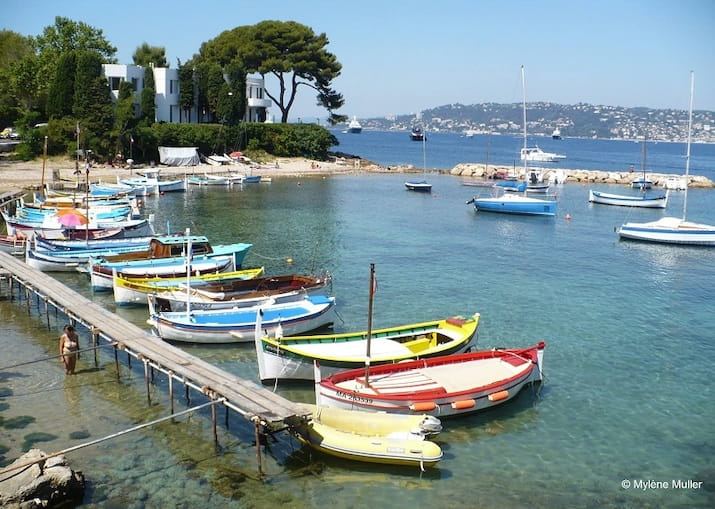 Colorful boats moored at a wooden dock by a clear, calm sea with distant hills.