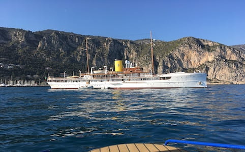 Large white yacht with yellow funnel near rocky coastline under blue sky.
