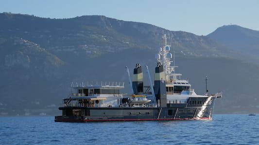 Large yacht with two smokestacks on a calm sea, mountains in background.