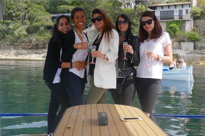 Five women on a boat holding glasses with water view in background.
