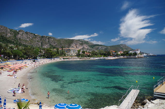 Sunny beach scene with clear blue water, mountains, and people relaxing.