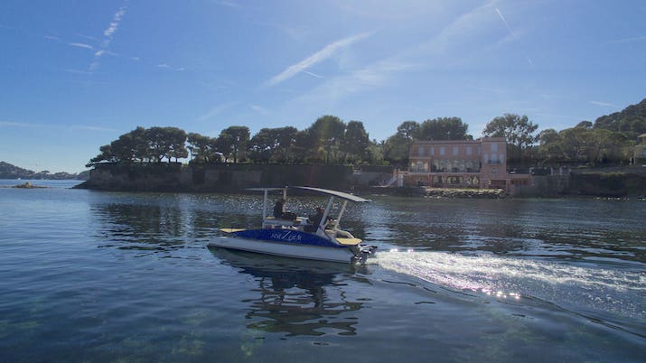 Solar-powered boat on calm water near a shoreline with trees and buildings.