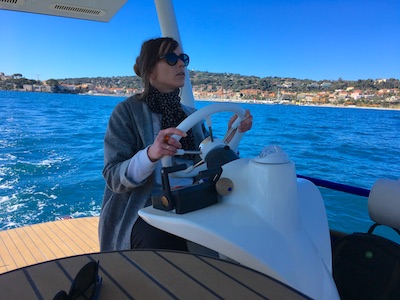 Woman steering a boat on a sunny day with a coastline in the background.