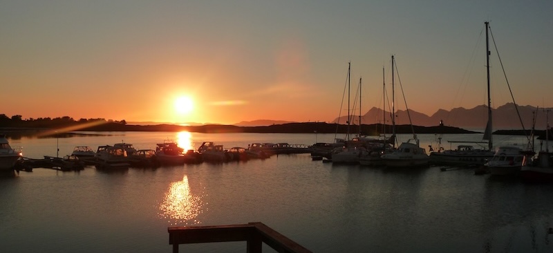 Sunset over a calm harbor with anchored boats and distant mountains.