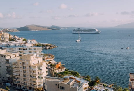 Coastal cityscape with buildings, cruise ship, and sailboat on a calm sea.
