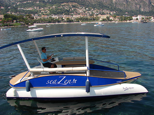 Man on a solar-powered boat with canopy floats on a scenic lake.