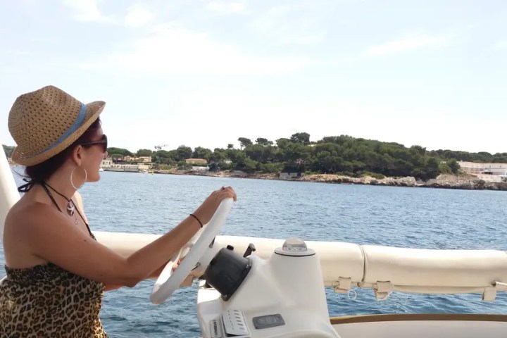 Woman in hat steering a boat on a sunny day, with coastline visible in the background.