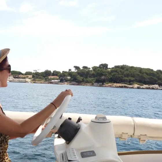 Woman in hat steering a boat on a sunny day, with coastline visible in the background.