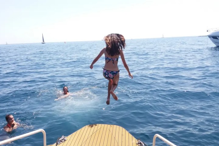 Girl in bikini jumps off boat into the sea, with two people swimming nearby.