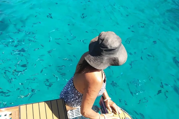 Woman in a swimsuit and hat sits on a boat deck by clear blue water with small fish.