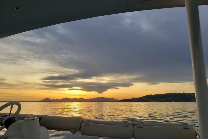 View from boat at sunset over calm sea with distant hills and cloudy sky.