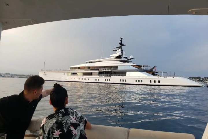 Two people admire a large white yacht on calm water under a clear sky.