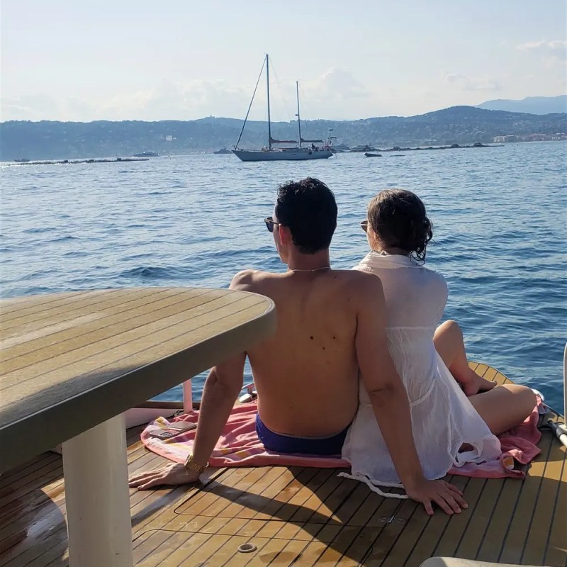 Couple sitting on a boat deck, gazing at the sea with a sailboat in the distance.