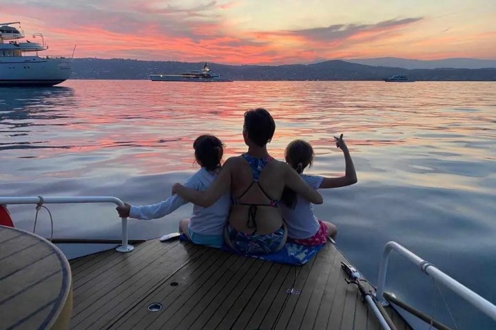 Three people sit on a boat deck watching a colorful sunset over calm waters.