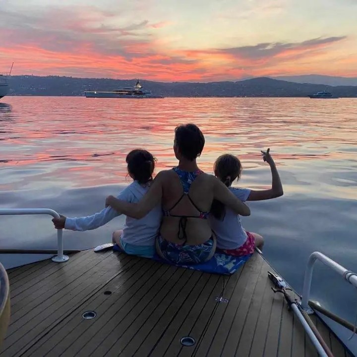 Three people sit on a boat deck watching a colorful sunset over calm waters.