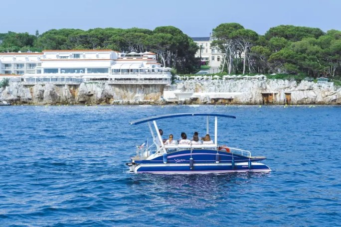 Small boat with people on blue water, rocky shoreline and buildings in background.