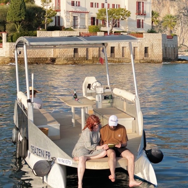 Two people sitting on the edge of a boat in calm water near a villa.