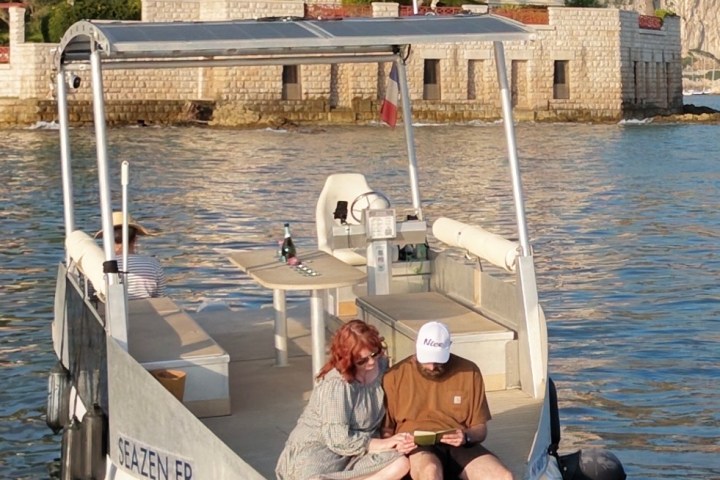 Two people sitting on the edge of a boat in calm water near a villa.