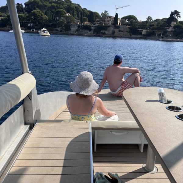 Two people relaxing on a boat deck by the water, with trees and buildings in the background.