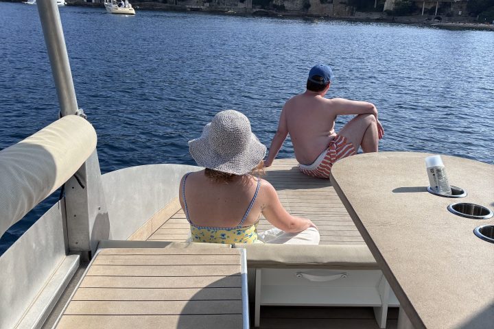 Two people relaxing on a boat deck by the water, with trees and buildings in the background.