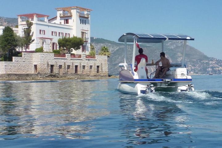 Two people on a small motorboat near a waterfront house with mountains in the background.