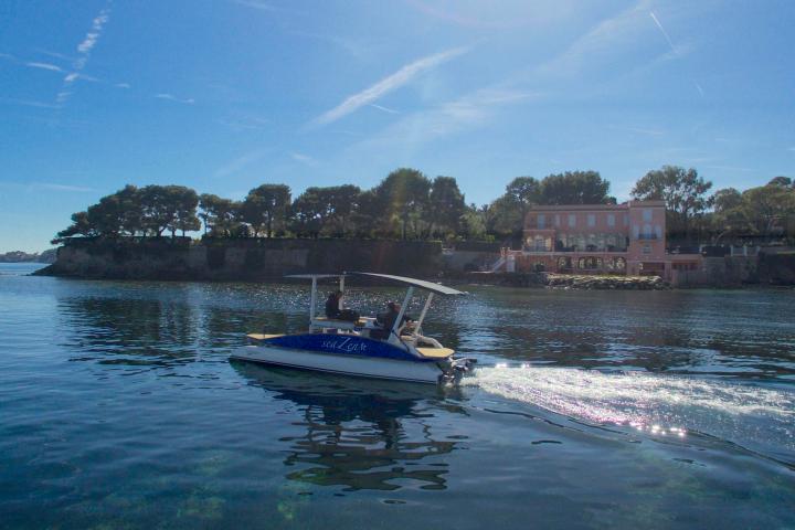 Small boat on calm water near a pink building and trees under a clear blue sky.