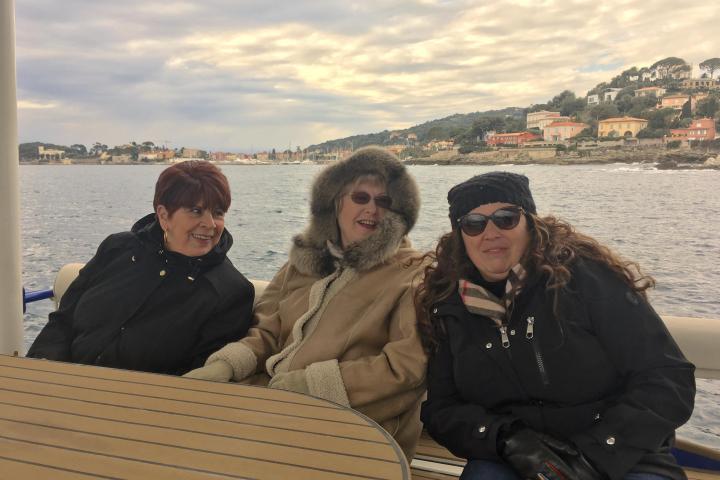 Three women in winter clothes sit on a boat with a coastal view in the background.