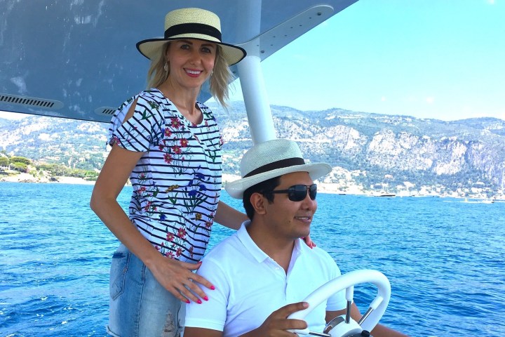 Two people wearing hats on a boat with mountains and sea in the background.