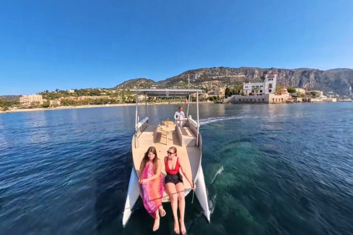Two women relaxing on a boat with a coastal town and mountains in the background under a clear blue sky.