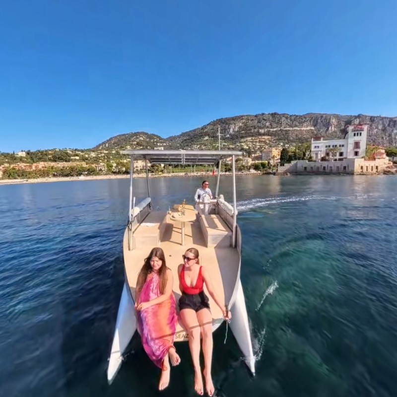 Two women relaxing on a boat with a coastal town and mountains in the background under a clear blue sky.