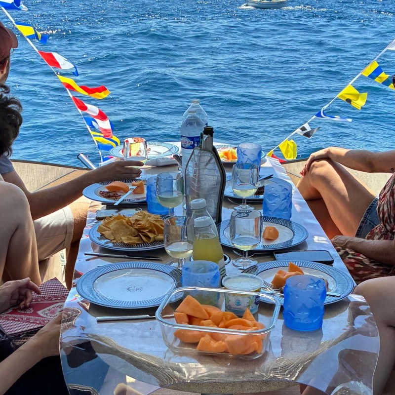 Five people on a boat with a table of snacks and drinks, colorful flags overhead, and ocean in the background.