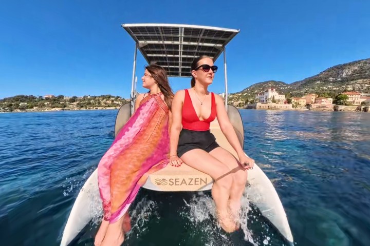 Two women sitting on a solar-powered boat enjoying a sunny day on the water with hills and buildings in the background.
