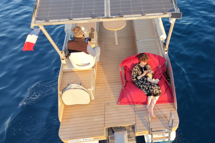Two people on a solar-powered boat; one relaxing on a bean bag near water.
