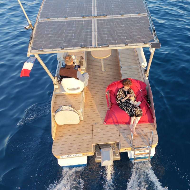 Two people on a solar-powered boat; one relaxing on a bean bag near water.