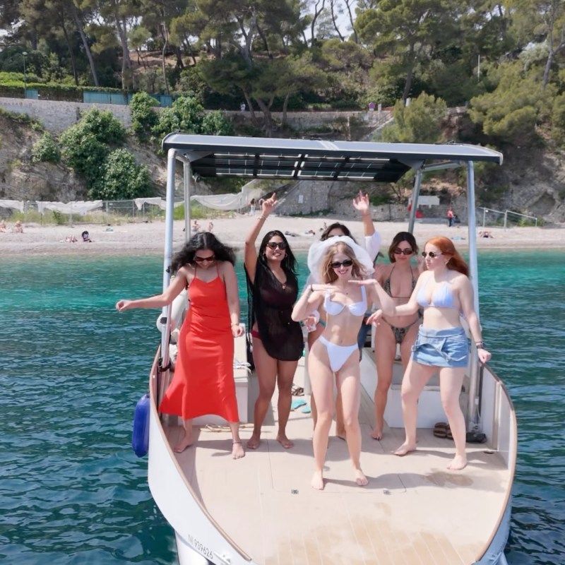 Six people on a boat, enjoying sunny weather near a beach.