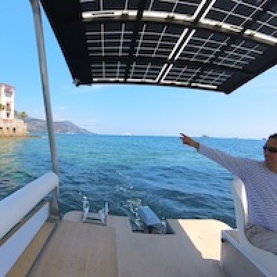 Person on boat pointing towards the sea with a building and hills in the background.