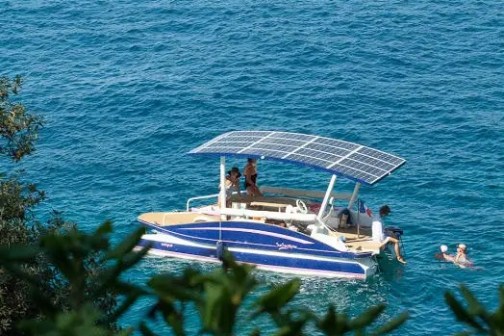 Boat with solar panels on blue water, people swimming nearby, trees in foreground.