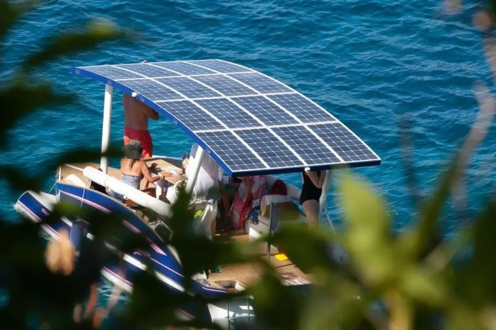 People on boat with solar panel roof on clear blue water, leaves in foreground.