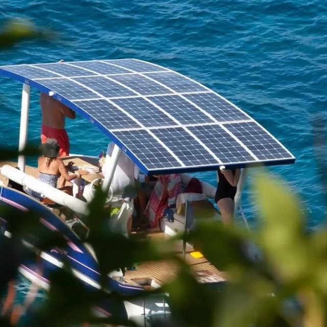 People on boat with solar panel roof on clear blue water, leaves in foreground.