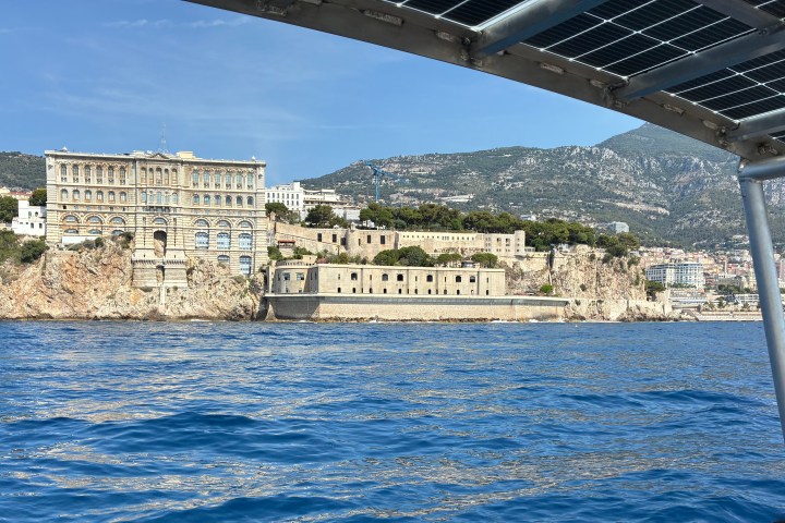 Coastal view of a grand building on a rocky cliff, seen from a boat with a solar panel canopy.