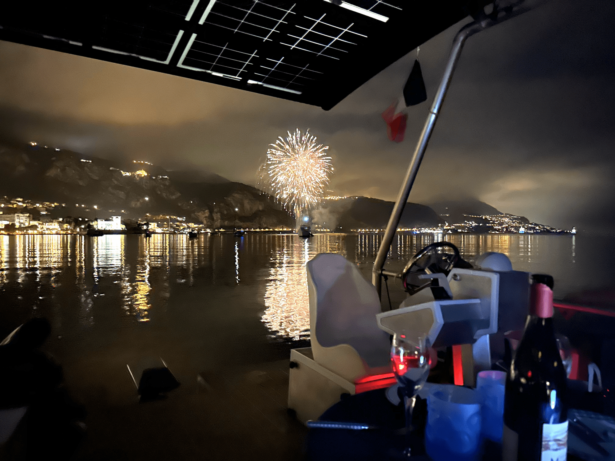Fireworks over a calm sea viewed from a boat at night, with mountains and city lights in the background.