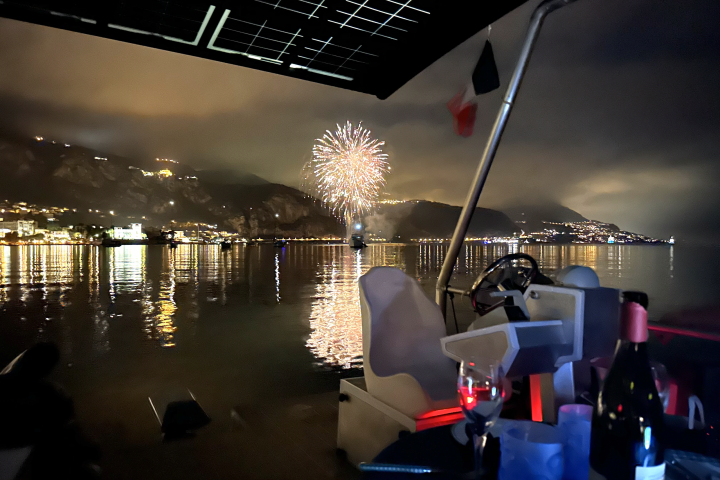 Fireworks over a calm sea viewed from a boat at night, with mountains and city lights in the background.